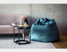 Modern eingerichtete Wohnzimmerecke mit grauem Sofa und blauem Sitzsack vor einer rohen Betonwand. Davor steht ein runder schwarzer Beistelltisch mit Büchern und einer Lampe auf einem hellen Teppich neben einem Holzfußboden.