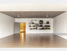 Minimalistischer Raum mit einem Holzboden, einer Beton-Decke und weißen Wänden. Rechts befindet sich ein Regal mit Yogamatten, Handtüchern, Pflanzen und Fitnessutensilien; links führt eine Tür in einen warm beleuchteten Bereich mit Sitzkissen.