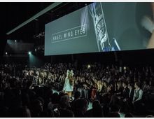 Eine Modenschau mit einem beleuchteten Laufsteg, umgeben von dicht besetzten Zuschauerreihen in einem dunklen Innenraum. Im Hintergrund hängt ein großer Bildschirm, der "ANGEL WING EYES" und ein Kosmetikprodukt zeigt.