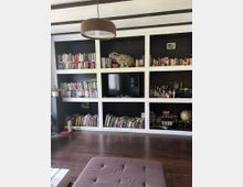 Wohnzimmer mit eingebauter Regalwand, die Bücher, Dekorationsgegenstände, eine TV-Anlage und andere Accessoires enthält. Der Raum hat dunklen Holzboden, eine gepolsterte Ottomane im Vordergrund und eine Deckenlampe in schlichtem Design.