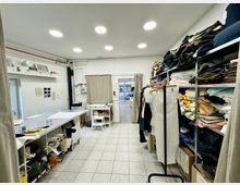 Ein Stoff- und Schneideratelier mit Regalen voller bunter Stoffe und Materialien auf der rechten Seite, einem großen Arbeitstisch links mit Nähmustern und Werkzeugen sowie einer kleinen Durchgangstür mit Vorhang im Hintergrund. Der Raum ist hell beleuchtet und hat einen gefliesten Boden.