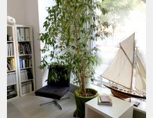 Ein heller Raum mit einem großen Fenster, einer hohen Zimmerpflanze, einem schwarzen Sessel und einem Modellsegelboot auf der Fensterbank. Links steht ein Bücherregal mit Büchern und Ordnern, und im Vordergrund befindet sich ein weißer Tisch mit Magazinen.