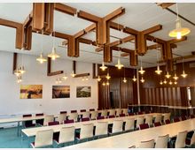 Ein Konferenzraum mit hellen Holztischen und hellen Stühlen, die teils mit weinroten Sitzpolstern versehen sind. Die Decke ist mit hölzernen Zierleisten dekoriert, von denen zahlreiche gelbe Hängelampen herabhängen; an der weißen Wand hängen drei Landschaftsfotografien.