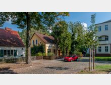 Straßenszene mit Kopfsteinpflaster, einer Reihe von kleinen Wohnhäusern mit Satteldächern und farbigen Fassaden. Im Vordergrund steht ein rotes Auto, umgeben von Bäumen und gepflegten Vorgärten.