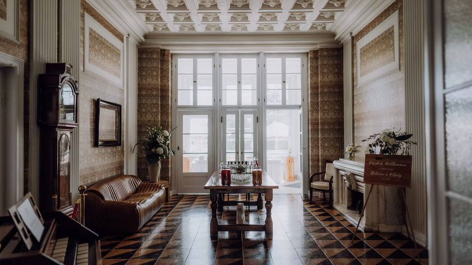 Elegante Halle im historischen Stil mit Kassettendecke, gemusterten Wänden und schwarz-braunem Fliesenboden. In der Mitte steht ein Holztisch mit Getränkeflaschen vor hohen verglasten Türen; seitlich sind ein Ledersofa, eine Standuhr sowie ein Kamin mit Blumendekoration zu sehen.