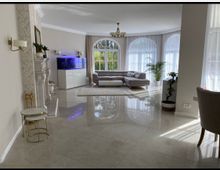 Helles Wohnzimmer mit großen, bodentiefen Fenstern, einem grauen Ecksofa, einem rechteckigen Couchtisch und mehreren Zimmerpflanzen. Im Raum befinden sich ein Kamin mit Dekoration, ein großes Aquarium und glänzende Fliesenboden.