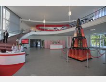 Geräumiger Eingangsbereich eines modernen Gebäudes mit einer geschwungenen Treppe, einem Empfangstresen, einer Garderobe und einer dekorativen Installation aus stapelbaren Elementen. Große Fenster lassen Tageslicht herein, und rote Details ziehen sich durch die Gestaltung.