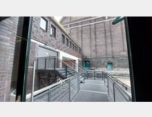 Blick auf einen Innenhof mit Backsteinfassaden, einer Außentreppe aus Metall und einer Metallrampe. Im Hintergrund ist eine massive Mauer mit grünen Türen erkennbar.