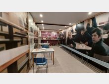 Ein Raum, der wie eine historische Schulmensa gestaltet ist, mit Tischen, Stühlen und einer langen Bank an den Wänden. Die Wände sind mit großformatigen Fotografien von Schülern und einer Schulszenen-Dekoration versehen, im Hintergrund ist ein Tresen mit Utensilien und Vorräten zu sehen.