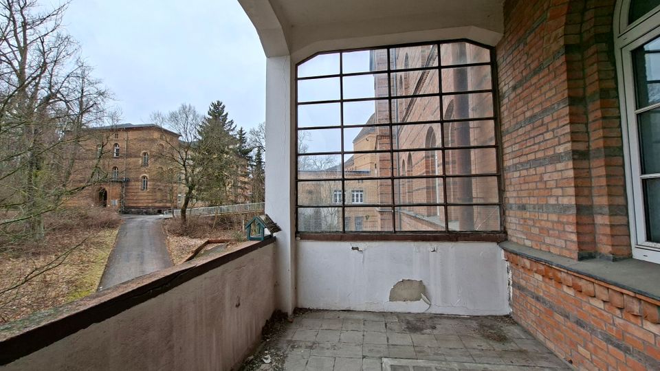 Überdachter Laubengang oder Balkon an einem Backsteingebäude mit Fliesenboden und einer großen, gitterartig gerahmten Fensterfront. Links führt ein Weg durch kahlen Baumbestand zu weiteren Backsteinbauten im Hof.