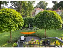 Ein gepflegter Garten mit zwei kleinen, kugelförmigen Bäumen, zwischen denen eine orangefarbene Hängematte gespannt ist. Im Vordergrund befindet sich eine Terrasse mit schwarzen Gartenmöbeln und blühenden Pflanzen; im Hintergrund sind Hecken, Blumenbeete und Wohnhäuser sichtbar.