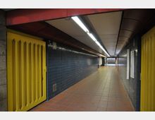 Ein unterirdischer Fußgängertunnel mit orangefarbenen Fliesenboden und dunkelblau gefliesten Wänden. Gelbe Türen befinden sich auf beiden Seiten, und die Decke ist mit langen, linearen Leuchtstofflampen ausgestattet. Im Hintergrund führen Treppen nach oben.