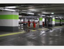 Unterirdisches Parkhaus mit Betonwänden, die durch grüne und graue Streifen hervorgehoben sind, und mehreren roten Schranken zur Zufahrt. Die nassen Bodenflächen reflektieren die Deckenbeleuchtung, und im Hintergrund sind neonbeleuchtete Notausgangsschilder sichtbar.