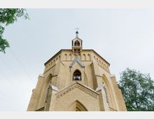 Außenansicht einer historischen Kirche aus gelbem Backstein mit Spitzbögen und einer Glockenöffnung im Turm. Im Hintergrund sind ein bewölkter Himmel und Baumkronen sichtbar.