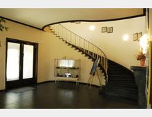 Ein Innenraum mit einer geschwungenen Treppe aus dunklem Holz und einem Geländer aus Metall an einer cremefarbenen Wand. Im Vordergrund steht eine Vitrine mit Ausstellungsstücken und eine Staffelei mit einem Gemälde, daneben eine zweiflügelige Glastür.