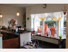 Ein kleines Café mit einer Theke aus Holz und Metall, darauf Flaschen, Gläser und eine Kaffeemaschine. Große Fenster mit weiß-gelben Vorhängen blicken auf gestapelte orangefarbene Stühle und eine Hecke draußen.