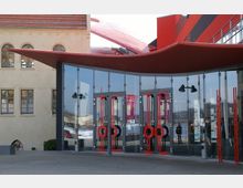 Moderner Eingangsbereich eines Theaters mit einer Fassade aus Glas und auffälligen roten Türelementen. Im Hintergrund ist ein helles historisches Gebäude und ein rotes Dach mit geschwungenen Formen zu sehen.