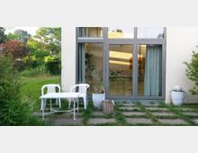 Außenansicht eines modernen Gebäudes mit großen, bodentiefen Fenstern. Davor befinden sich ein weißer Plastiktisch mit zwei Stühlen, ein kleiner Garten mit Steinplatten und Gras sowie dekorative Pflanzen in Töpfen.