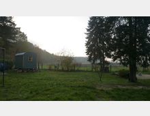 Grüne Wiese am Waldrand mit einem kleinen, dunkelgrünen Bauwagen mit Fenster links, umgeben von Sträuchern und einem Zaun. Im Hintergrund liegen offene Felder, rechts stehen hohe Nadelbäume und ein gepflasterter Bereich.