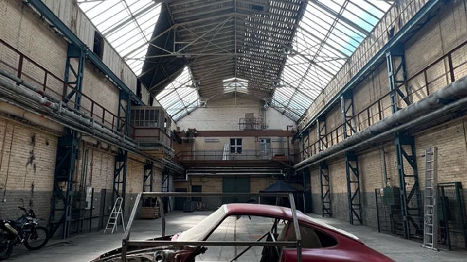 Große Industriehalle mit hohem Glasdach und sichtbaren Stahlträgern, flankiert von gemauerten Seitenwänden mit Rohrleitungen und Laufstegen. In der Mitte steht ein teilweise zerlegter Autokarosserie-Rohbau auf einem Metallgestell auf dem Betonboden.