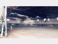 Verlassene Industriehalle mit hohen Betonwänden, mehreren kleinen Fenstern und einer Dachkonstruktion aus freiliegenden Stahlträgern. Der Boden ist mit Schmutz bedeckt, und die Wände zeigen Spuren von Graffiti.