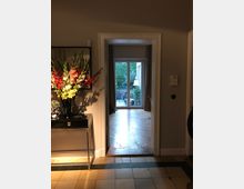 Flur in einem Wohnbereich mit Blick durch eine Tür auf einen Raum mit Parkettboden und einer verglasten Terrassentür, die zu einem Garten führt. Links im Flur steht ein Tisch mit einer Vase voller bunter Blumen.