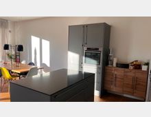 Moderne Küche mit grauen Einbaumöbeln, einem Herd und einem schwarzen Küchenblock in der Mitte. Neben der Küche steht ein Essbereich mit einem Holztisch, bunten Stühlen und hängenden schwarzen Lampen, im Hintergrund scheint Sonnenlicht durch ein Fenster.