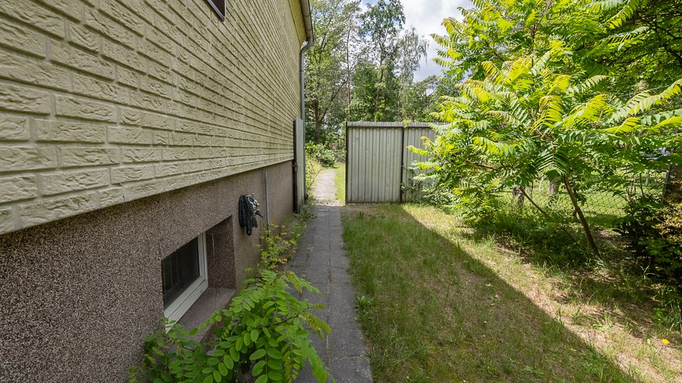 Schmaler Seitenbereich neben einem Wohnhaus mit hell verklinkerter Fassade und kleinem Kellerfenster, entlang eines gepflasterten Weges. Rechts grenzt eine grasbewachsene Fläche mit dichtem Strauch- und Baumbewuchs an, im Hintergrund steht ein graues Metalltor oder Schuppen vor einem Waldsaum.