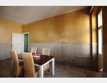 Ein einfacher, heruntergekommener Raum mit gelblich-braun gestreifter Tapete und einer halbhoch grauen Wandverkleidung. In der Mitte steht ein Tisch mit vier geflochtenen Stühlen, ein offenes weißes Türblatt und ein Fenster lassen Tageslicht herein.