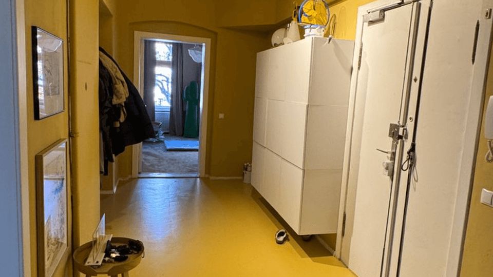 Long, narrow interior hallway with yellow walls and floor, exposed wooden beams overhead, and a doorway at the far end leading to a brighter room. White storage cabinets and a large white door line the right wall, with framed pictures and a small stool along the left side.