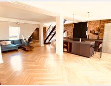 Großzügiger, moderner Wohnraum mit offenem Grundriss, bestehend aus einer Küche mit dunkelgrauer Kochinsel, einer Ziegelsteinwand, einer hölzernen Treppe und einem Wohnzimmer mit blauer Eckcouch und Couchtisch. Der Boden ist aus hellem Holz im Fischgrätmuster verlegt, und die Räume sind durch große Fenster gut beleuchtet.