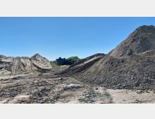 Erde- und Schutthügel auf einer Baustelle mit klar erkennbarem, unebenem Boden, tiefen Reifenspuren und Maschinenteilen im Hintergrund vor einem strahlend blauen Himmel.