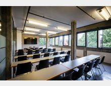 Moderner Seminarraum mit rechteckigen Tischen und schwarzen Stühlen, ausgerichtet in Reihen. Große Fenster auf der rechten Seite bieten Blick auf Bäume, während eine große Wand mit einem Flachbildschirm und einer Flipchart vorne sichtbar ist.