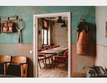 Raum mit pastellgrünen Wänden, an denen ein Lederrucksack, ein Tennisschläger und ein Boxsack hängen; durch eine offene Tür ist ein rustikaler Besprechungsraum mit Holztisch, roten Stühlen und großen Fenstern zu sehen.