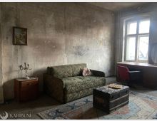 Abgenutztes Wohnzimmer mit stark verschmutzten, unverputzten Wänden, einem gemusterten Sofa und einem kleinen Beistelltisch mit Kerzenleuchter. Neben einem großen Fenster stehen ein Schreibtisch mit Stuhl; auf einem Teppich liegt eine große Holztruhe als Couchtisch.