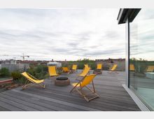 Rooftop-Terrasse mit Holzboden, gelben Liegestühlen und kleinen runden Beistelltischen, umgeben von einer Glasbrüstung mit Blick auf städtische Gebäude, Bäume und einen bewölkten Himmel.
