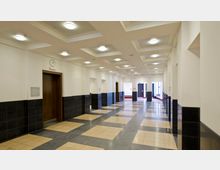 Ein heller Flur in einem modernen Gebäude mit einem Schachbrettmuster aus beigen und schwarzen Bodenfliesen. Die Wände sind in Weiß gehalten, mit schwarzen Fließen im unteren Bereich; an der Decke befinden sich rechteckige Vertiefungen mit runden Deckenleuchten, und am Ende des Flurs sind Türen und Fenster erkennbar.