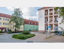 Mehrstöckige Wohnhäuser in Pastellfarben, teils mit Balkonen und roten Ziegeldächern, umgeben eine kleine, bepflanzte Verkehrsinsel. Im Vordergrund befindet sich ein überdachter Fahrradständer neben einem Straßenschild der Ernst-Thälmann-Straße.