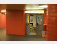 Ein Imbisslokal mit der Leuchtschrift „INTERNATIONALER SCHNELLIMBISS“ in einer Passage mit roten Fliesenwänden. Die offene Tür gewährt einen Blick auf den Innenraum mit weißen Kacheln, einfachen Holzmöbeln und einem Müllbehälter neben der Tür.