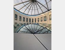 Innenansicht eines modernen Gebäudes mit kuppelartigem Glasdach und einem kreisförmigen Atrium. Die Wände haben vertikale Fenster, und die Glaskonstruktion spiegelt sich in der darunterliegenden Fläche.