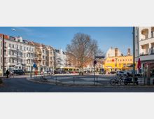 Straßenkreuzung in einer städtischen Umgebung mit mehrstöckigen Wohngebäuden im Gründerzeitstil auf der linken Seite, einem großen gelben Gebäude mit der Aufschrift „brotfabrik“ rechts sowie mehreren geparkten Autos. In der Mitte steht ein einzelner Baum, umgeben von Gehwegen und Radfahrern.