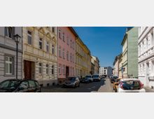 Straßenszene in einem Wohngebiet mit mehrstöckigen, farbigen Altbauten in Beige, Rosa und Grün; die gepflasterte Straße ist von geparkten Autos gesäumt und von einer Laterne beleuchtet, unter einem klaren blauen Himmel.