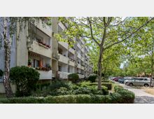 Wohngebäude mit hellen Balkonen, die mit Blumen dekoriert sind, umgeben von gepflegten Grünanlagen mit Büschen und Bäumen. Rechts im Bild verläuft ein Gehweg entlang einer kleinen Parkfläche, neben der Autos geparkt sind.