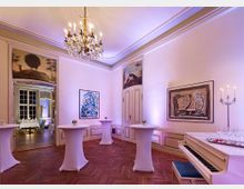 Eleganter Raum mit hellen Wänden, verziert mit Gemälden und historischen Wandmotiven. Es stehen weiße Stehtische mit Blumenarrangements auf einem Parkettboden, im Vordergrund ein weißes Klavier neben Kerzenständern, und im Hintergrund führt eine Tür in einen festlich gedeckten Essbereich.