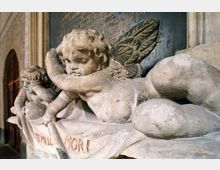 Steinrelief mit zwei detaillierten Engelfiguren auf einer Schrifttafel; einer der Engel liegt ausgestreckt mit Flügeln und hält ein Banner, auf dem „Memento Mori“ steht. Im Hintergrund sind goldene Inschrift und architektonische Details wie eine Tür und Wände sichtbar.