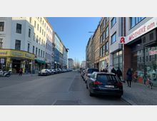 Straßenszene in einer Stadt mit mehrstöckigen Gebäuden auf beiden Seiten, darunter Geschäfte wie eine Apotheke und ein Restaurant. Geparkte Autos und Fußgänger befinden sich entlang der Gehwege, bei klarem Himmel und Tageslicht.