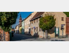 Eine schmale, gepflasterte Straße mit alten, teilweise renovierungsbedürftigen Häusern, von denen einige in Pastellfarben gestrichen sind. Im Hintergrund ragt ein Kirchturm in die Höhe, und rechts im Vordergrund stehen ein Baum, ein Touristeninformationsschild und eine Bank.