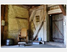 Innenraum eines traditionellen Fachwerkhauses mit sichtbaren, verwitterten Holzstützen und strohverputzten Wänden. Im Vordergrund stehen ein Schaukelstuhl, ein Polsterhocker und eine kleine Fußbank, links hängt eine kleine Laterne, rechts führt eine Holztür in einen anderen Raum.