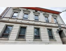 Fassade eines historischen Gebäudes mit Putz- und Mauerwerksschäden, verzierten Fensterrahmen im oberen Stockwerk und der Aufschrift „LPG Roter Stern Retzow Tierproduktion“ über den Erdgeschossfenstern. Rechts hängt eine Laterne neben einem Eingangsbereich.