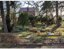 Gartenanlage mit großen Bäumen, Rasenflächen und Sträuchern sowie einem kleinen Teich mit Schilf am rechten Bildrand. Im Hintergrund steht ein Wohnhaus mit rotem Ziegeldach, teilweise durch das Gehölz verdeckt.